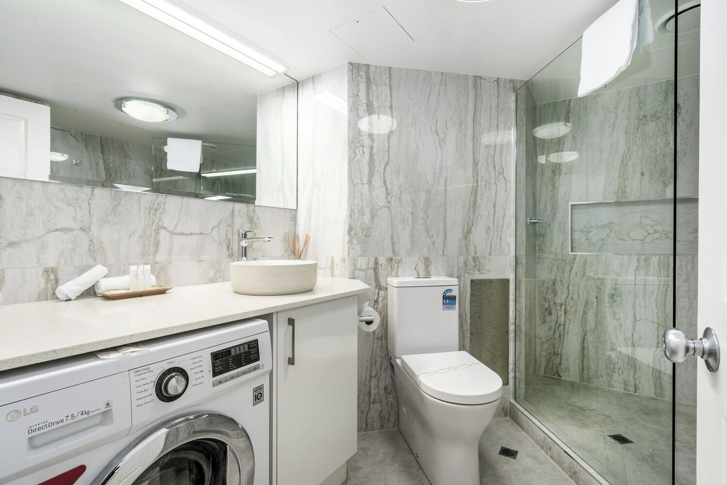 A modern bathroom with a marble backsplash, a built-in sink, a toilet, a washing machine, and a glass shower enclosure.