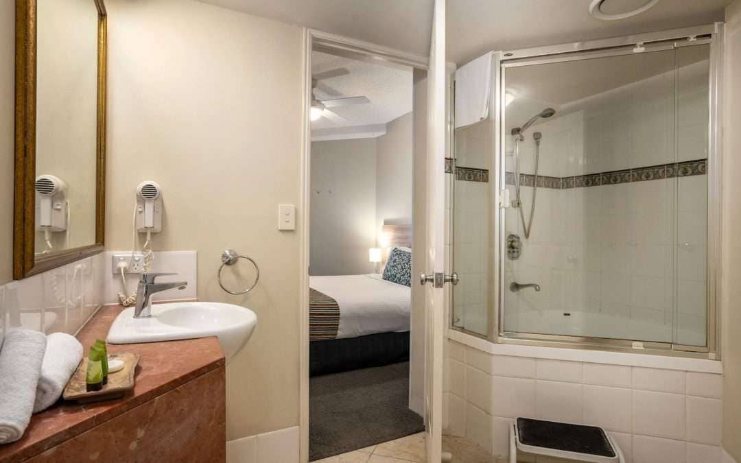 A hotel bathroom with a sink, mirror, hair dryers, a shower with glass doors, and a bed in the adjacent room.