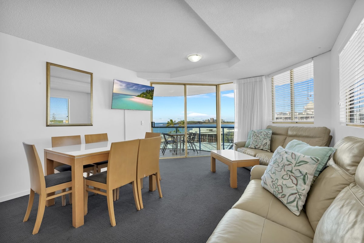 A room with a dining table, chairs, a sofa, a TV, and a balcony with a view of the sea.