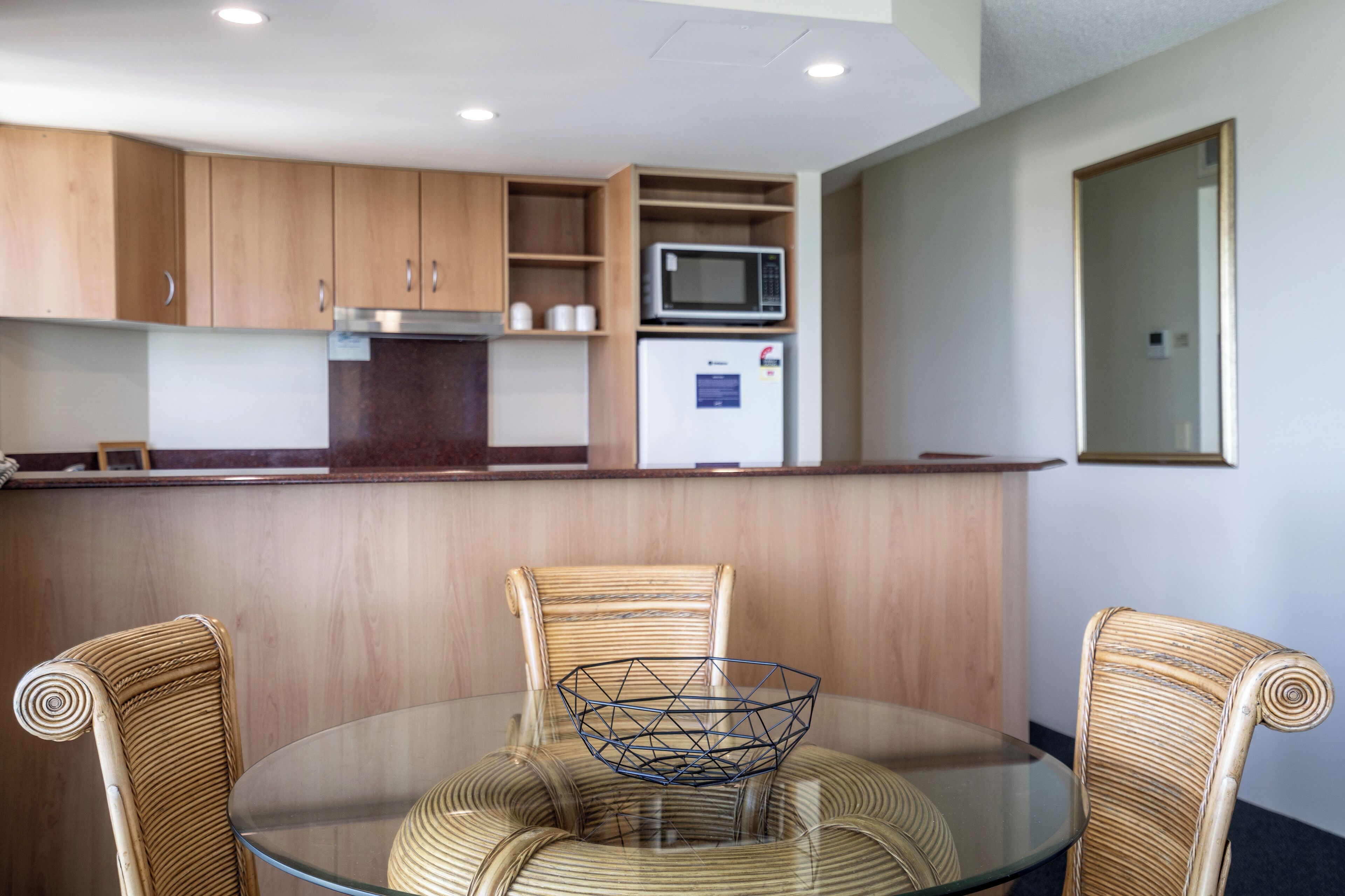 A modern kitchen with wooden cabinets, a microwave, and a refrigerator. There is a glass dining table with wicker chairs and a decorative glass bowl.