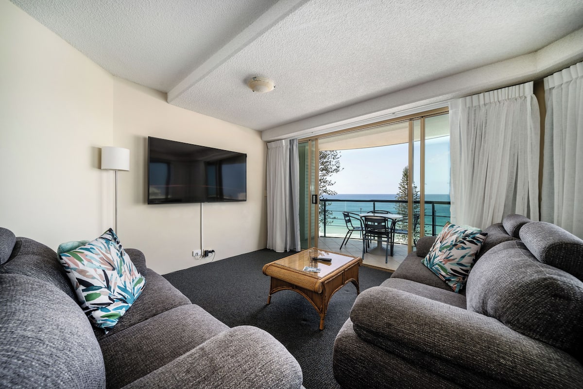 A modern living room with a flat-screen TV, grey sofas, a wooden coffee table, and a balcony with a view of the sea.