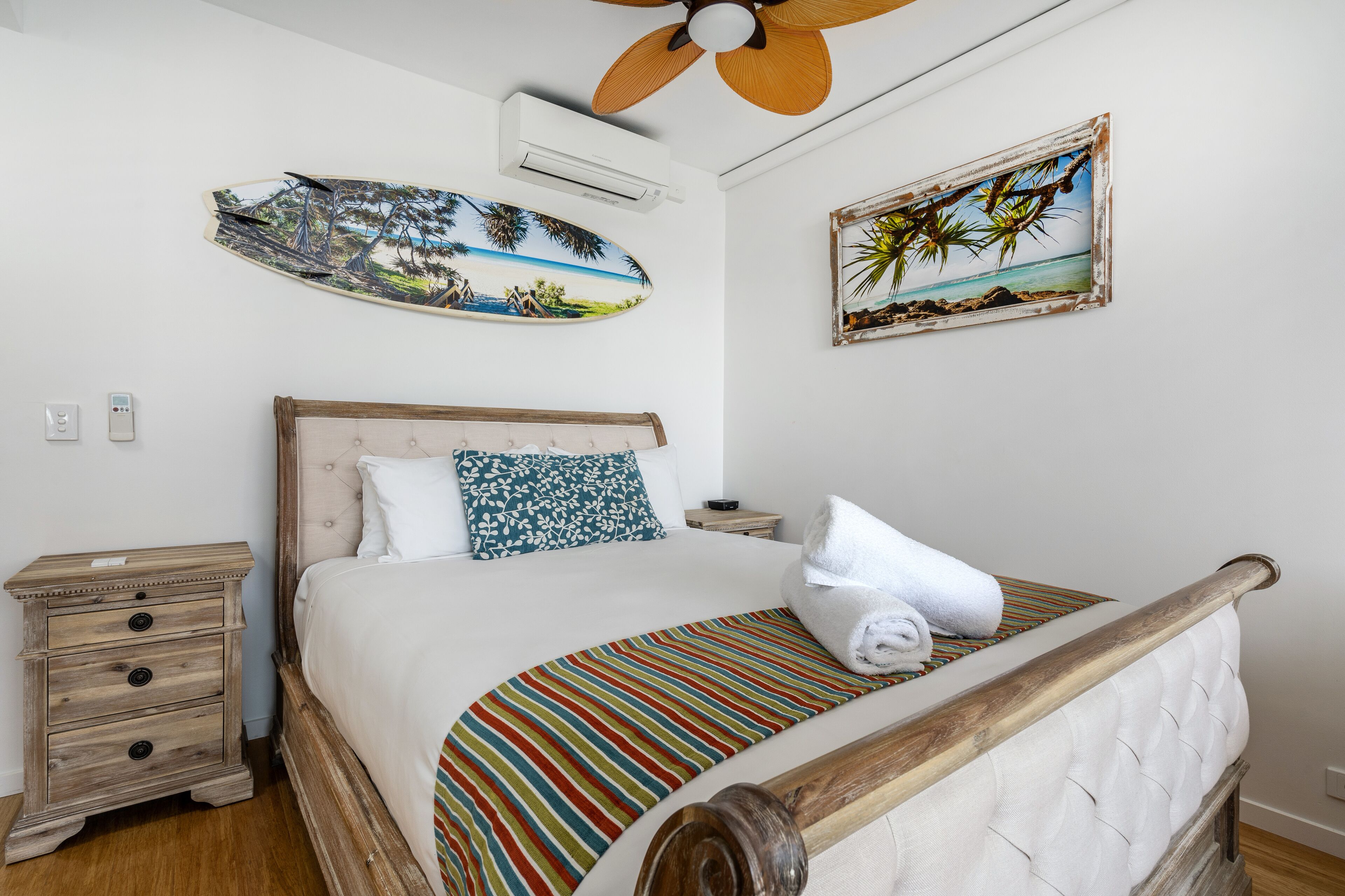 Quarto com estrutura de cama de madeira, colcha listrada, travesseiro branco, mesa de cabeceira com gavetas e ventilador de teto.