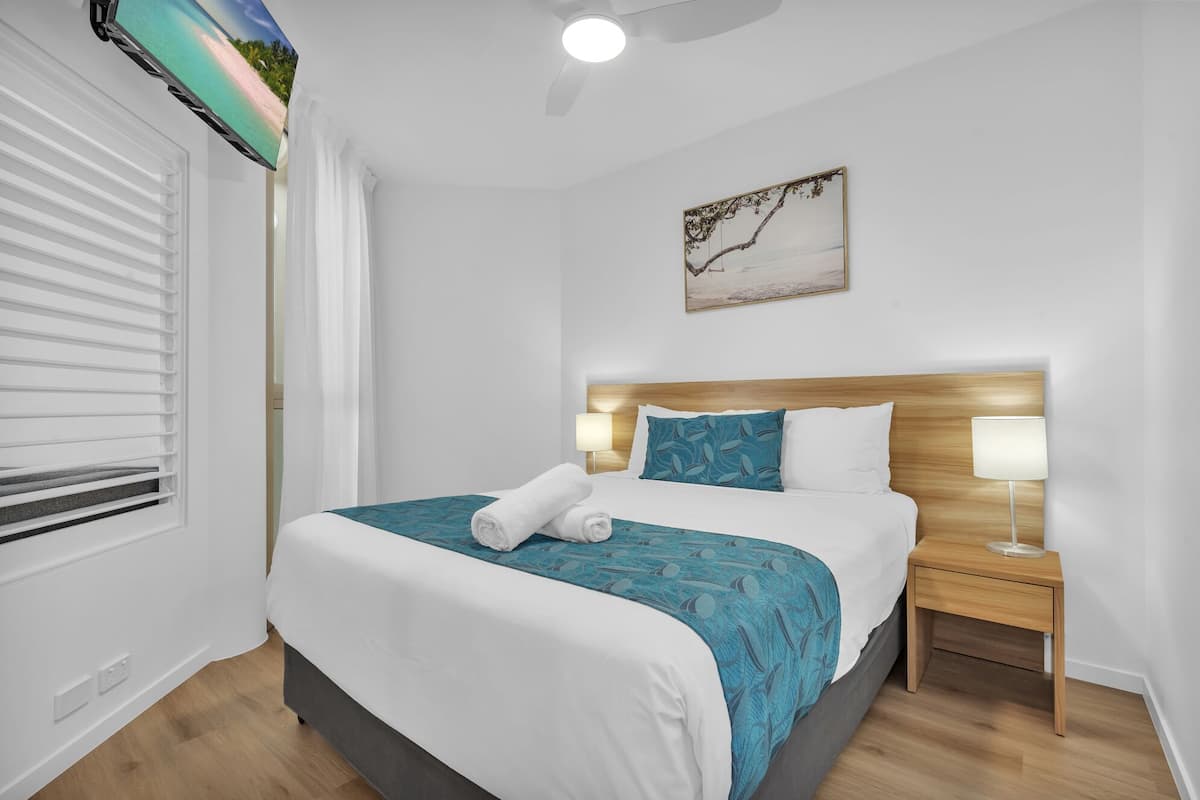 A hotel room with a bed, a nightstand, a lamp, a window with blinds, and a ceiling fan.