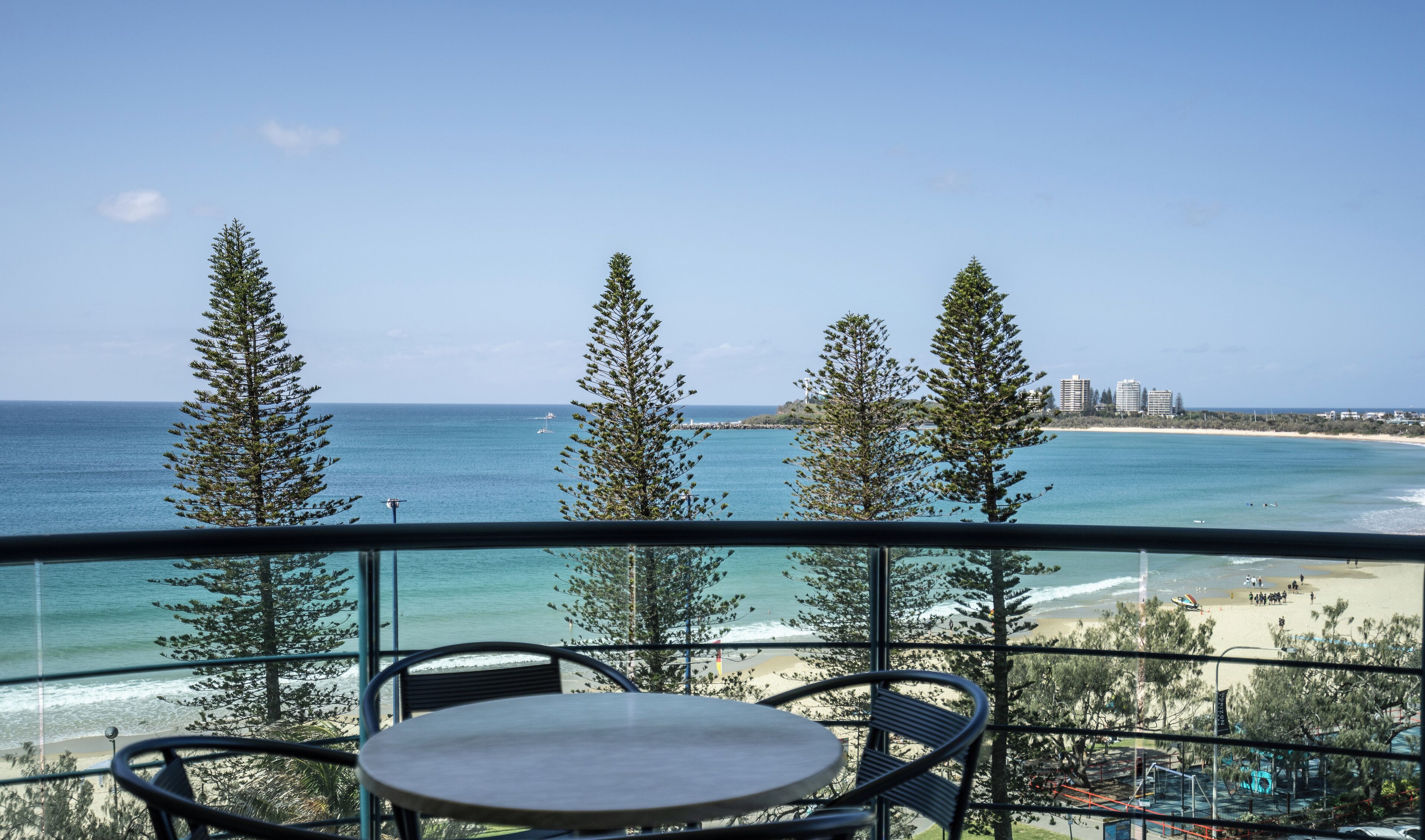 A balcony with a table and chairs overlooking a beach with trees and a clear sky.