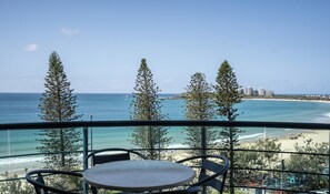 A balcony with a table and chairs overlooking a beach with trees and a clear sky.