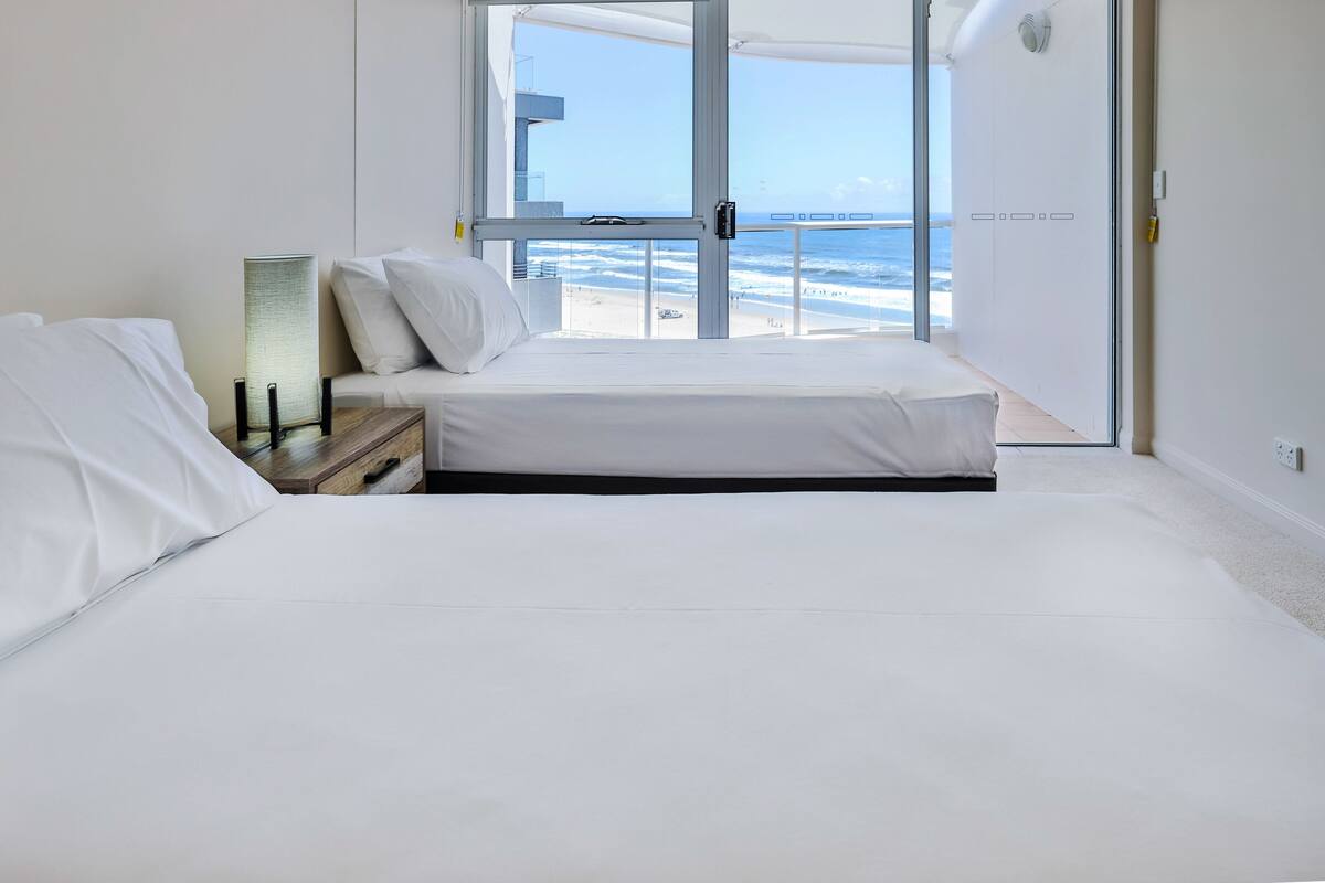 A bedroom with a bed, a bedside table, a lamp, and a view of the beach and ocean.