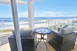 A balcony with wicker furniture and a glass table, overlooking a beach with waves. - Regency on the Beach (Palm Beach)