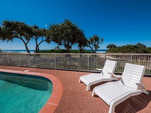 A pool area with two white lounge chairs and a view of the ocean.