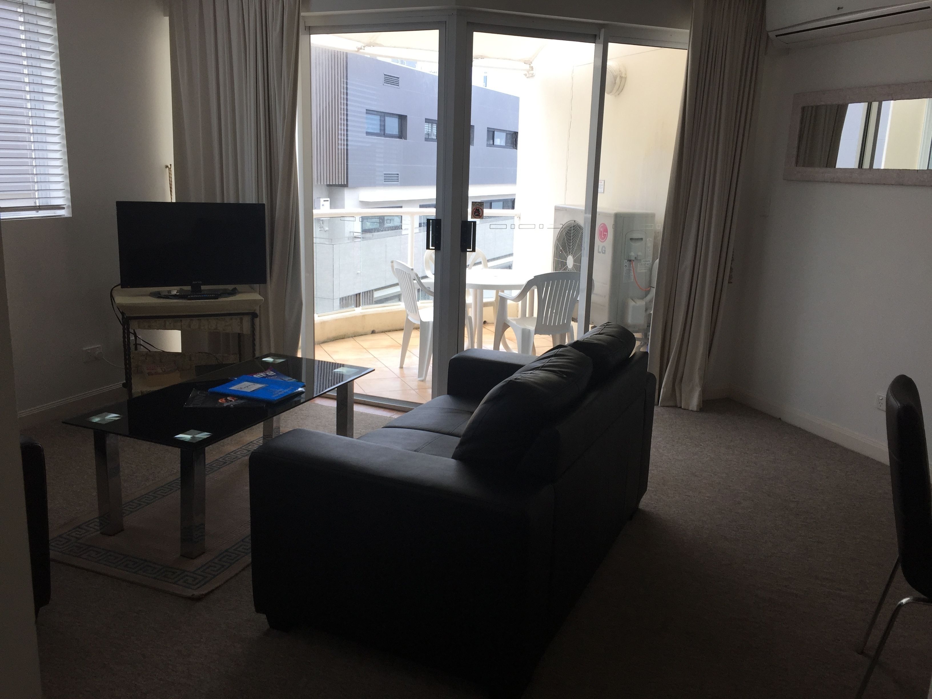 A living room with a black sofa, a glass coffee table, and a flat-screen TV. There is a sliding glass door leading to an outdoor area with a table and chairs.