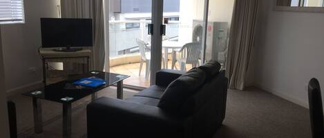 A living room with a black sofa, a glass coffee table, and a flat-screen TV. There is a sliding glass door leading to an outdoor area with a table and chairs.