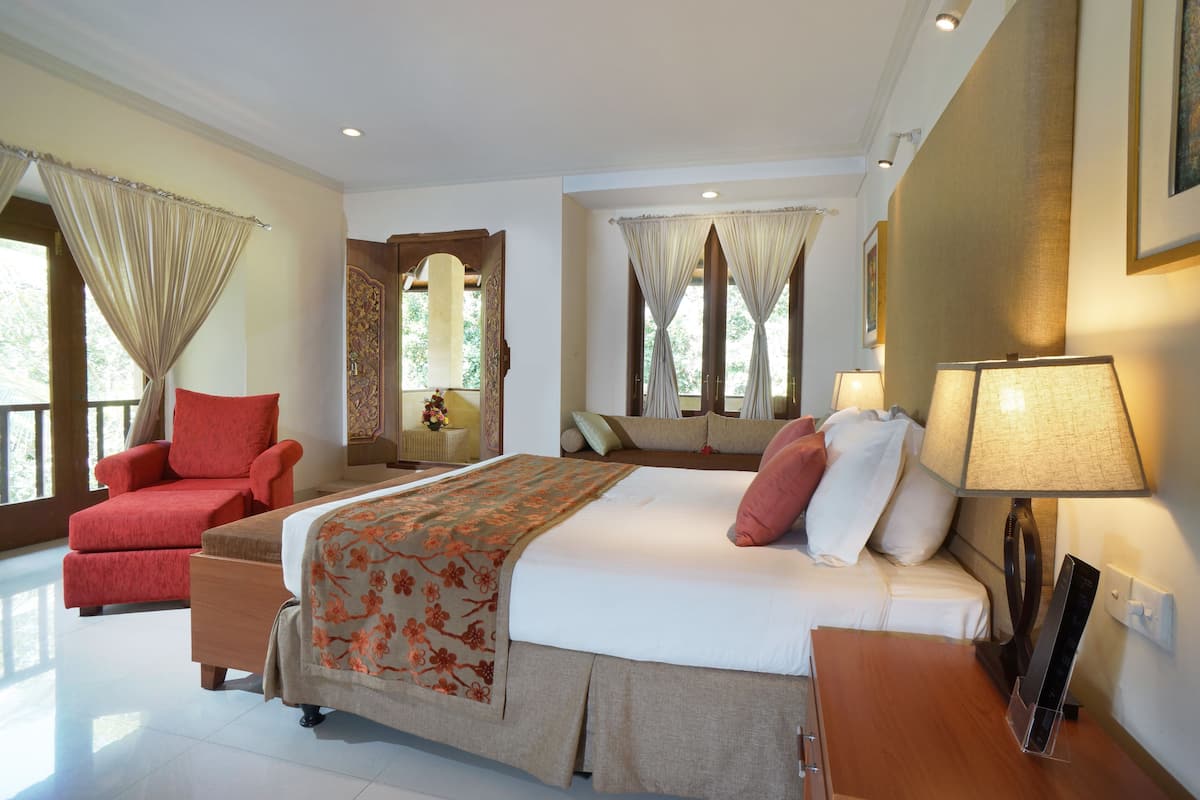 Habitación de hotel con una cama, un sillón rojo, una mesita de noche de madera, una lámpara y una ventana con cortinas.