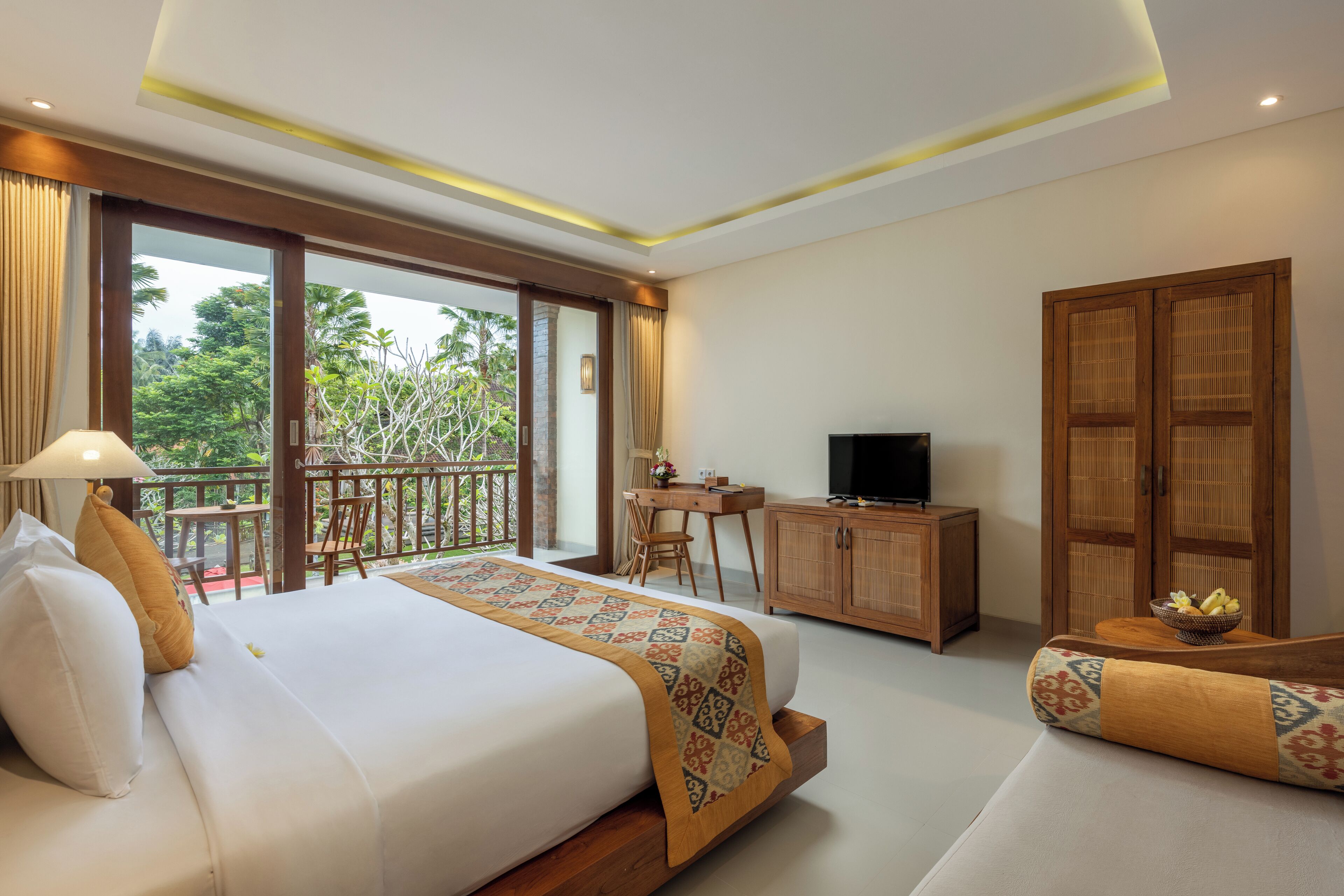 A hotel room with a bed, a desk, a television, and a balcony with a view of greenery.