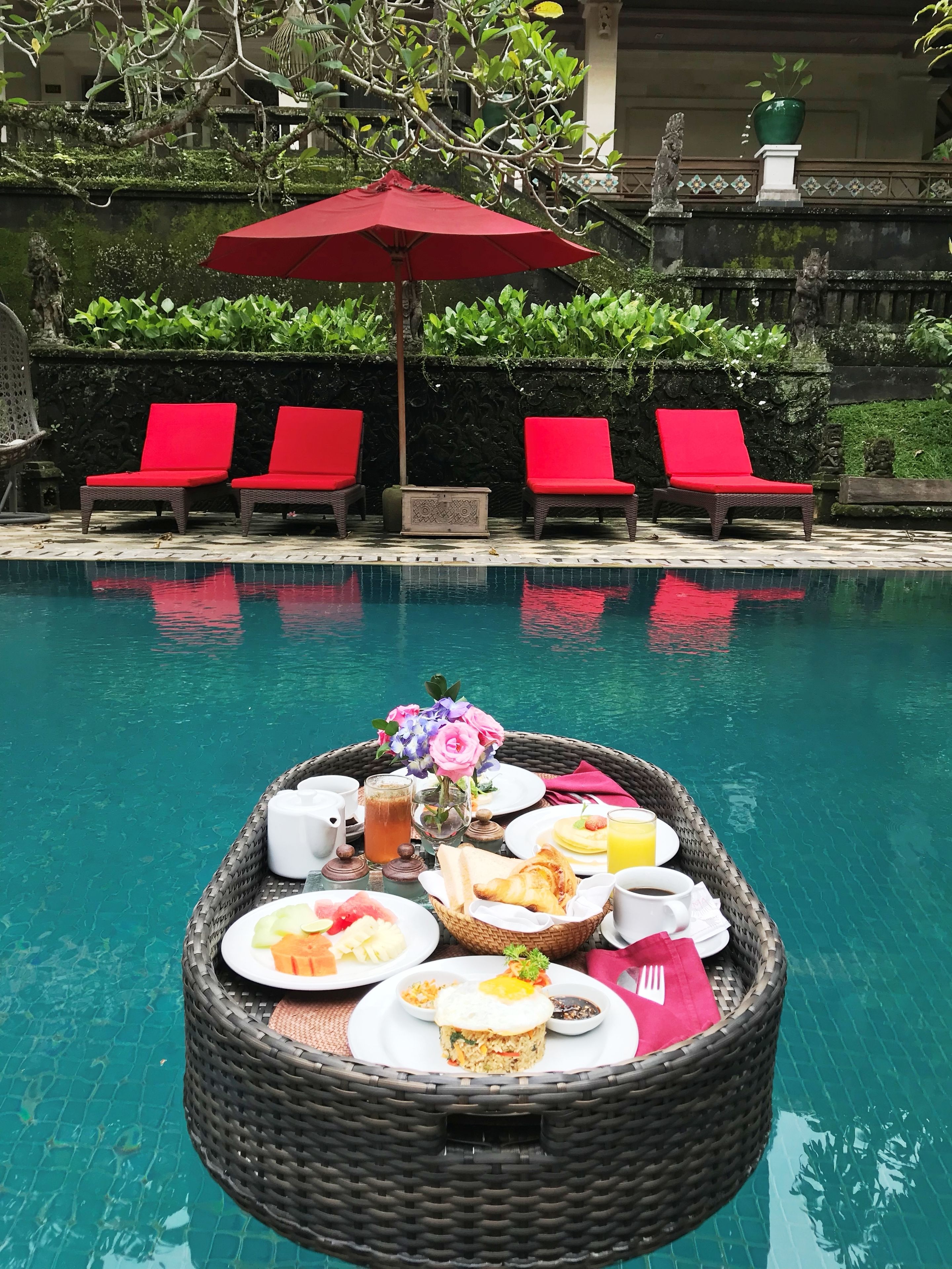 A poolside breakfast setup with a wicker basket containing plates of food, a drink, and a vase of flowers.