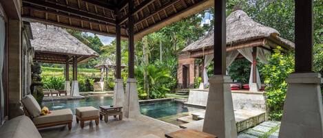 A covered outdoor area with lounge chairs, a pool, and thatched-roof structures.