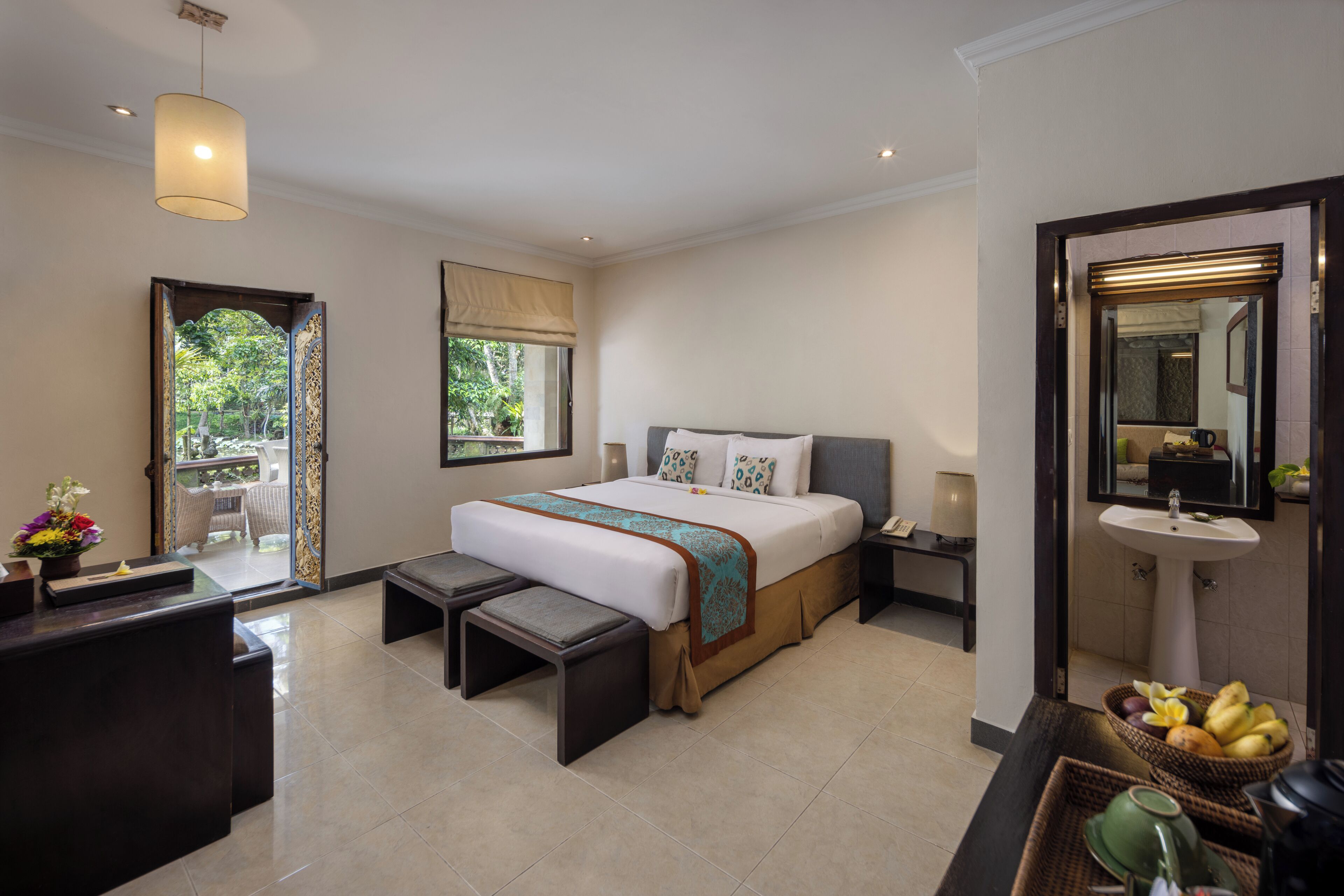 A hotel bedroom with a bed, bedside tables, a desk, a bench, a window, and a bathroom with a sink and mirror.