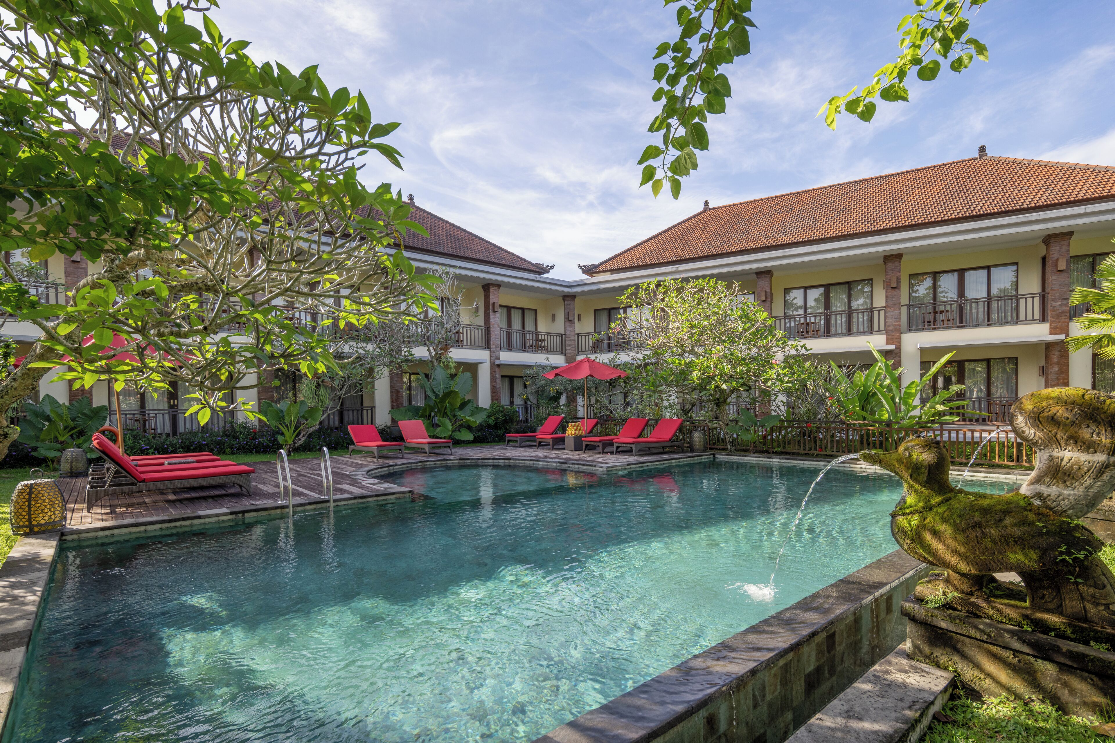 Ein Swimmingpool mit roten Liegestühlen, umgeben von einem Garten mit tropischen Pflanzen und einem Gebäude mit einem Ziegeldach.