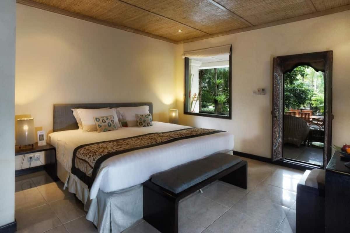 Un dormitorio con cama, mesitas de noche, un banco, una ventana y una puerta que da a un espacio exterior.