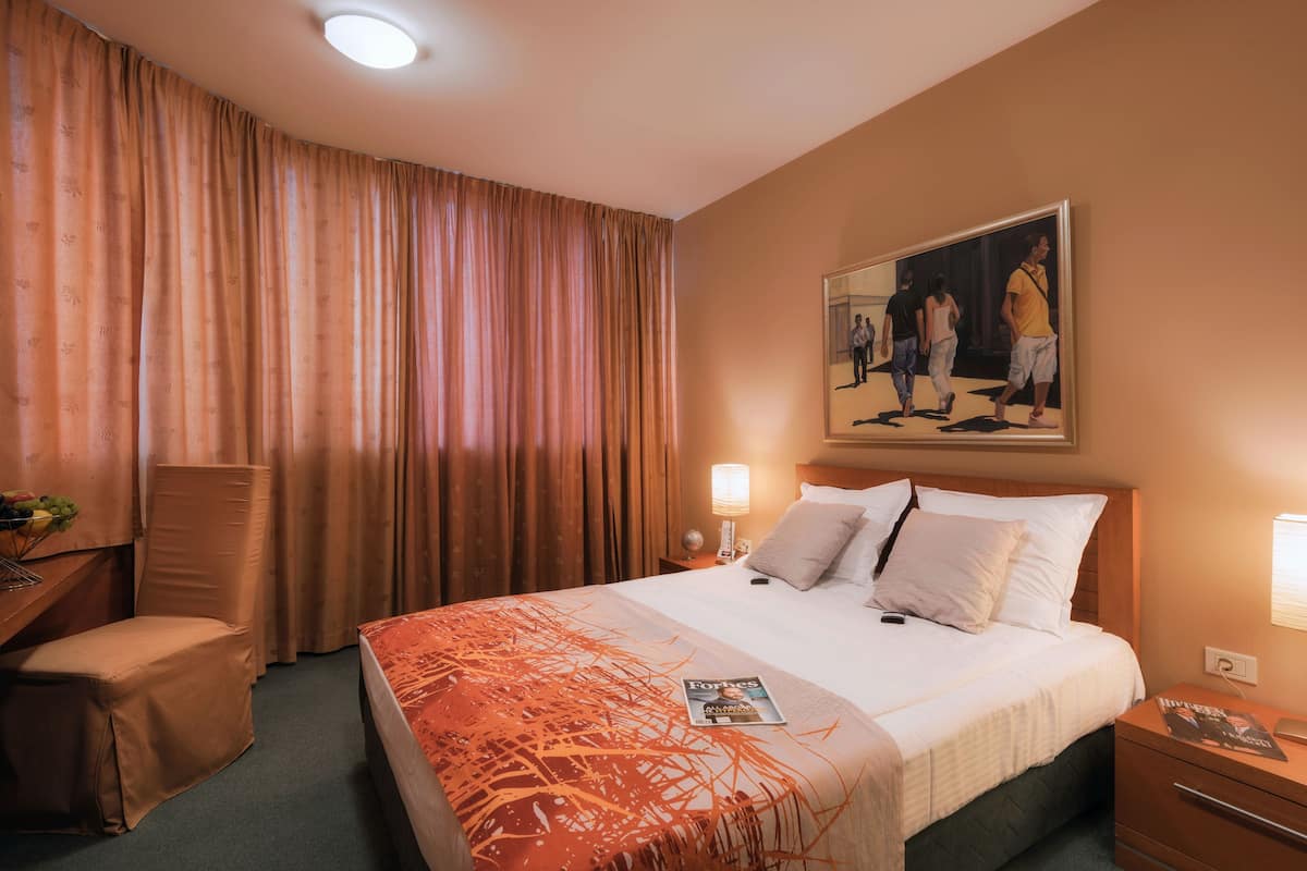 A hotel room with a large bed, a desk, a chair, a nightstand, and a painting on the wall.