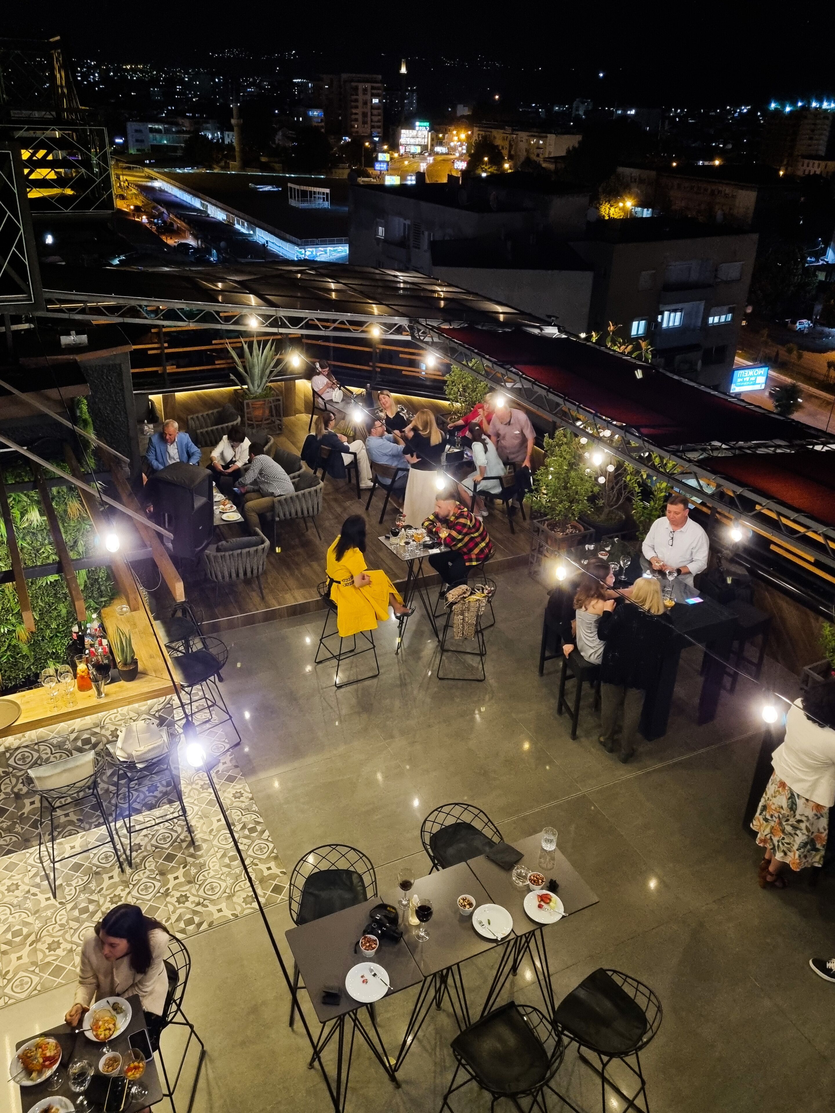 An outdoor dining area with tables and chairs, surrounded by plants and a cityscape view at night.