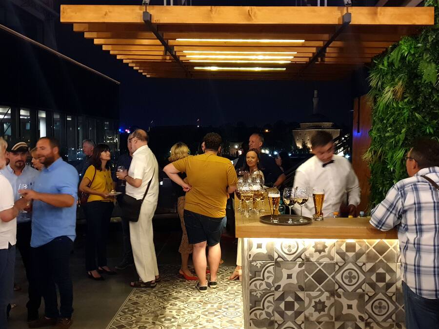 Evento social con gente de pie conversando en una zona de bar, con suelo de baldosas estampadas y techo de madera.