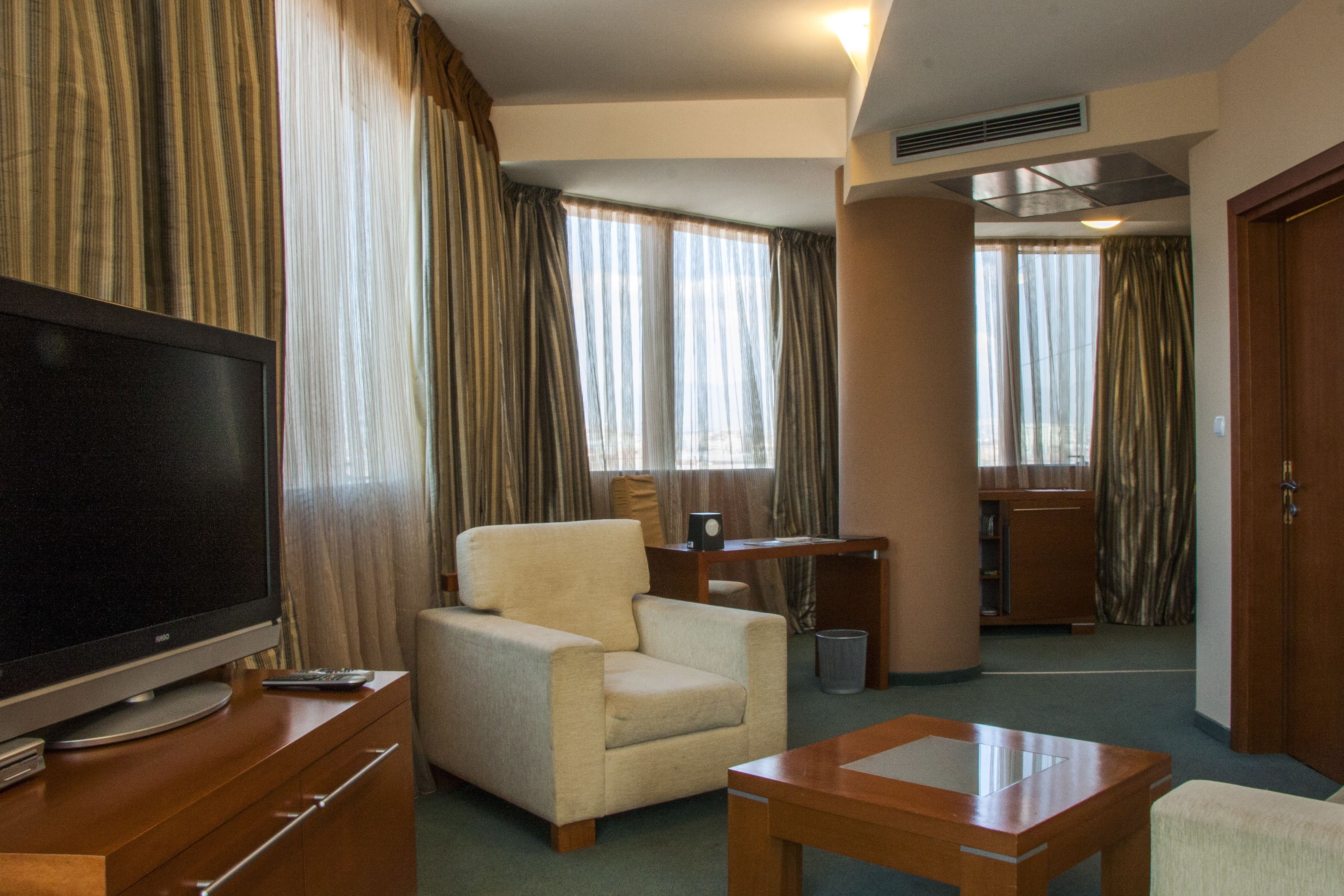 A hotel room with a television on a wooden stand, a beige armchair, a wooden table, and a desk with a chair.