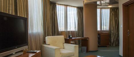 A hotel room with a television on a wooden stand, a beige armchair, a wooden table, and a desk with a chair.