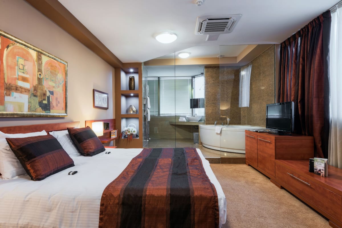 A hotel room with a large bed, a television, a desk, and a bathroom visible through a glass partition.