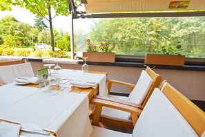 Un coin repas avec une table dressée pour deux personnes, des chaises en bois et une fenêtre donnant sur un espace verdoyant et une voiture garée à l’extérieur.