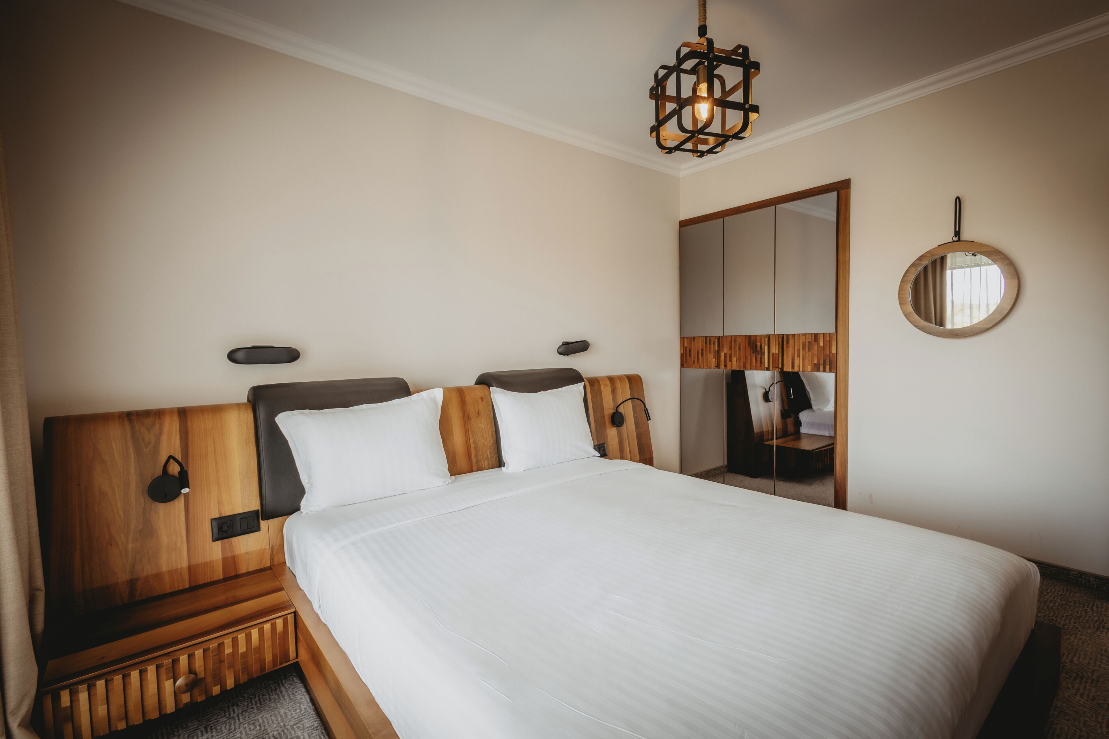 A neatly made bed with a wooden headboard, a bedside table, a wall-mounted mirror, and a hanging light fixture.
