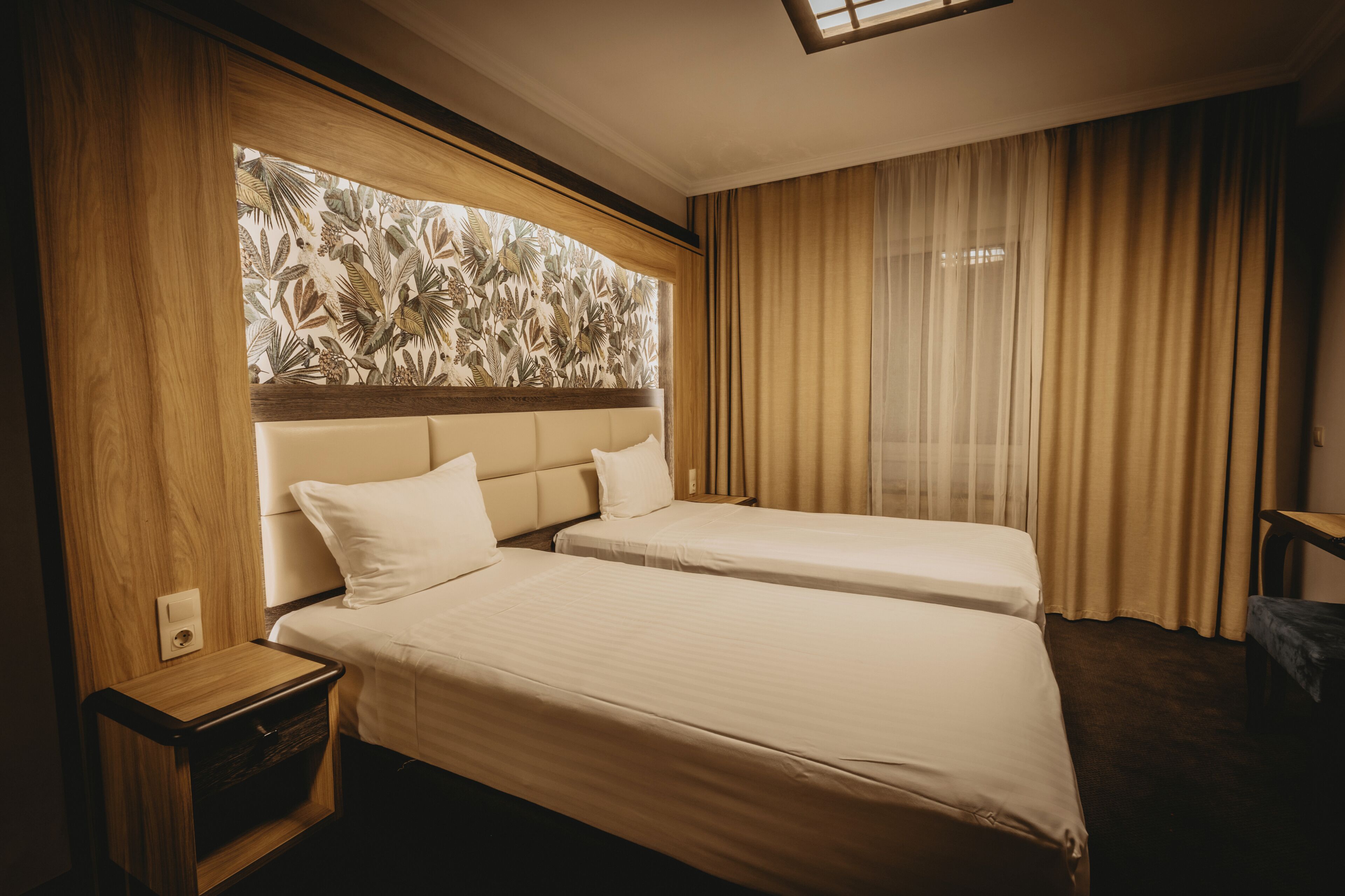 A hotel room with two beds, a wooden headboard, a nightstand, and a window with curtains.