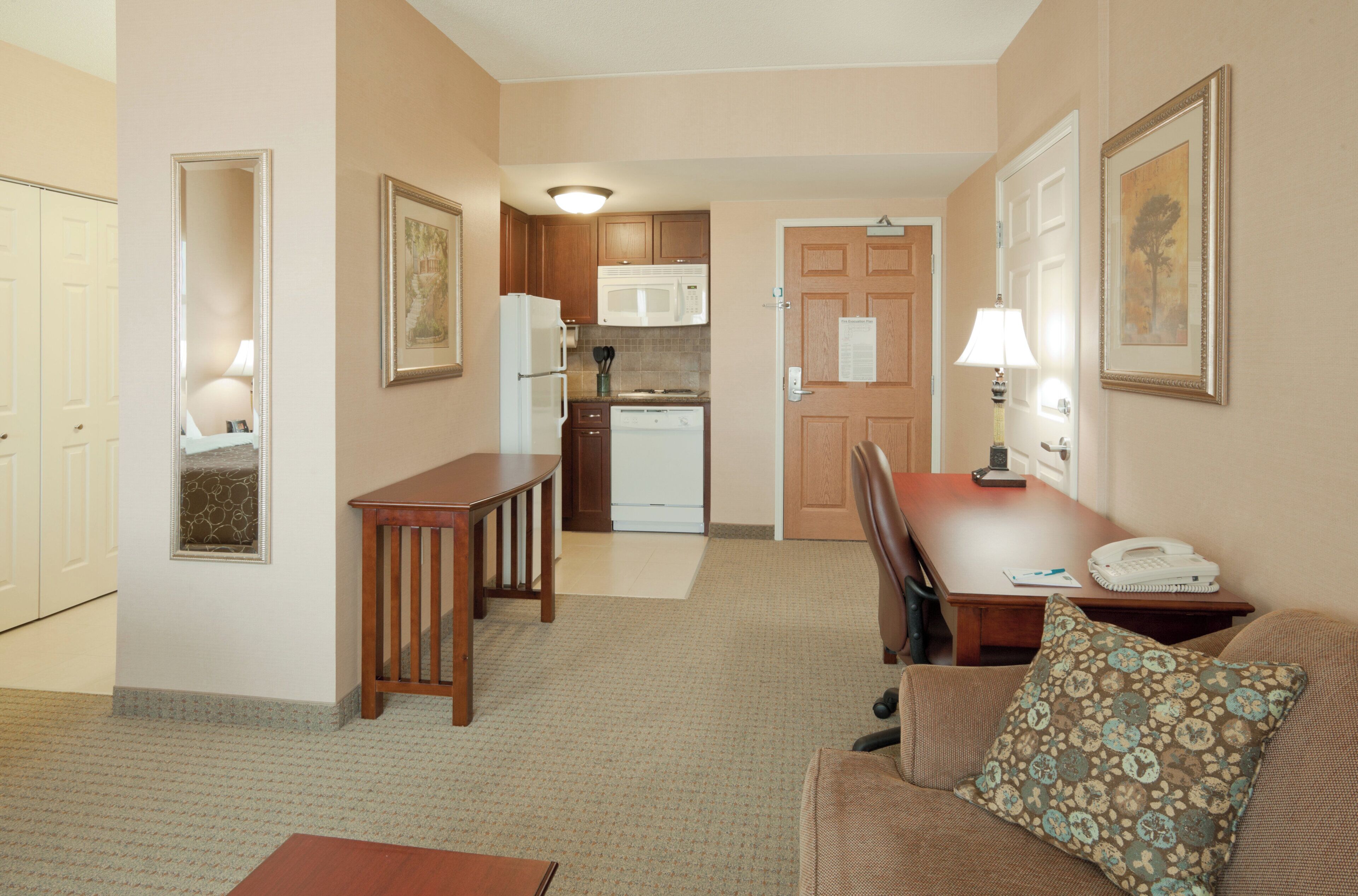 A compact hotel room with a kitchenette, a dining area with a table and chairs, and a living area with a sofa and a patterned pillow.