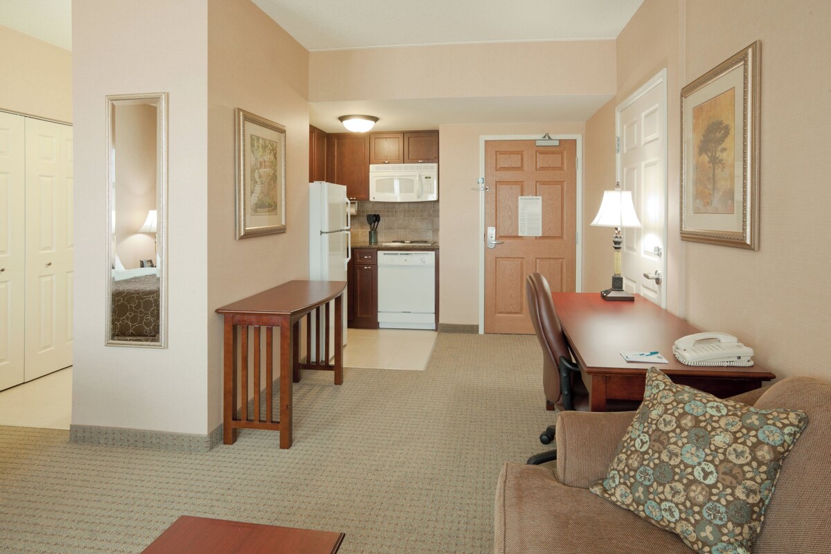 A compact hotel room with a kitchenette, a dining area with a table and chairs, and a living area with a sofa and a patterned pillow.