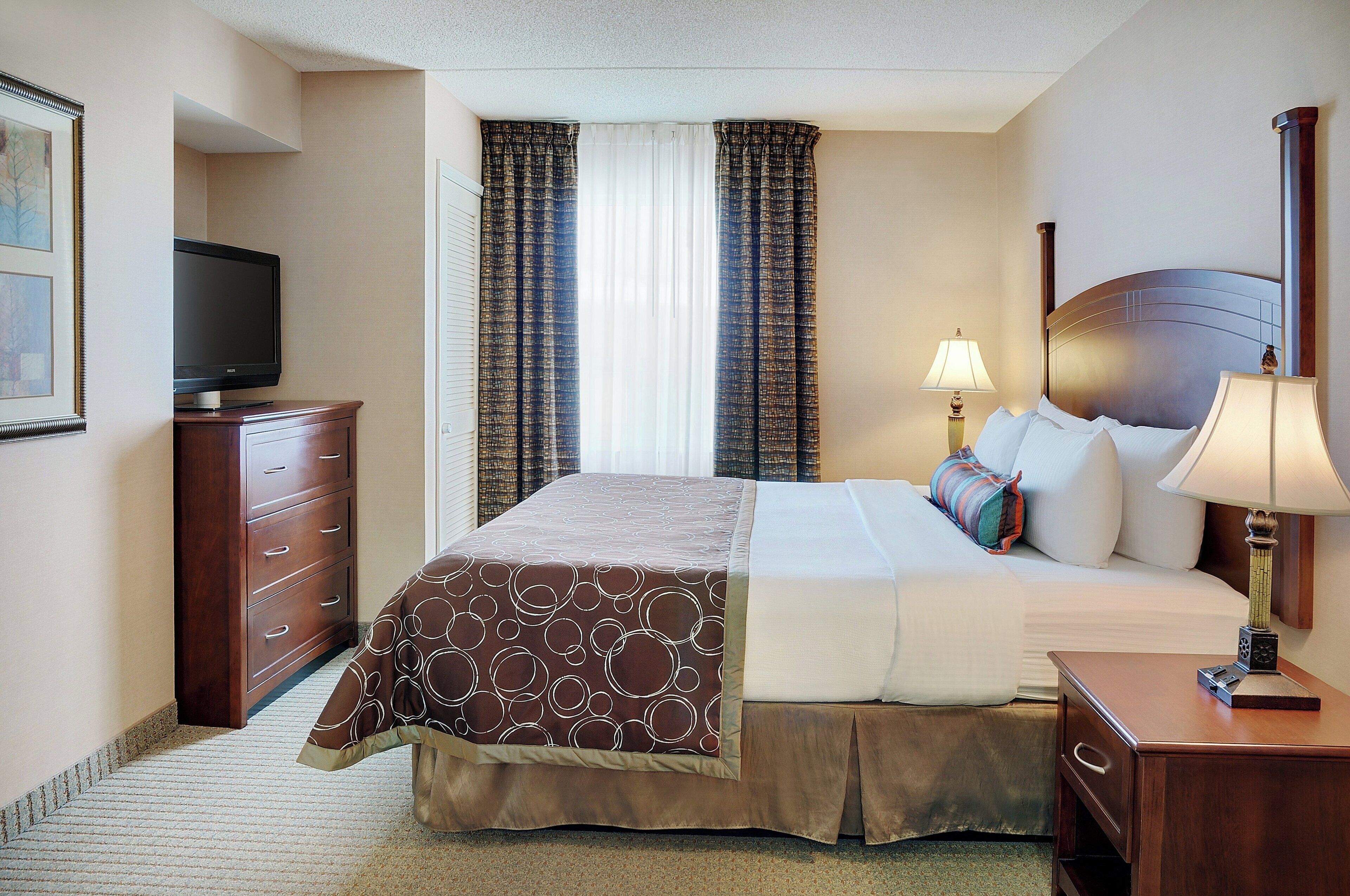 A hotel room with a bed, a television, a nightstand, a lamp, and a window with curtains.