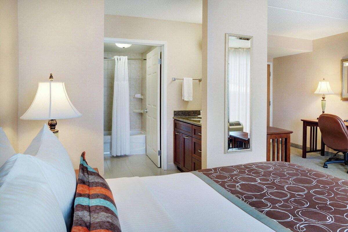 A hotel room with a bed, a desk, a chair, a bathroom with a shower, and a mirror.