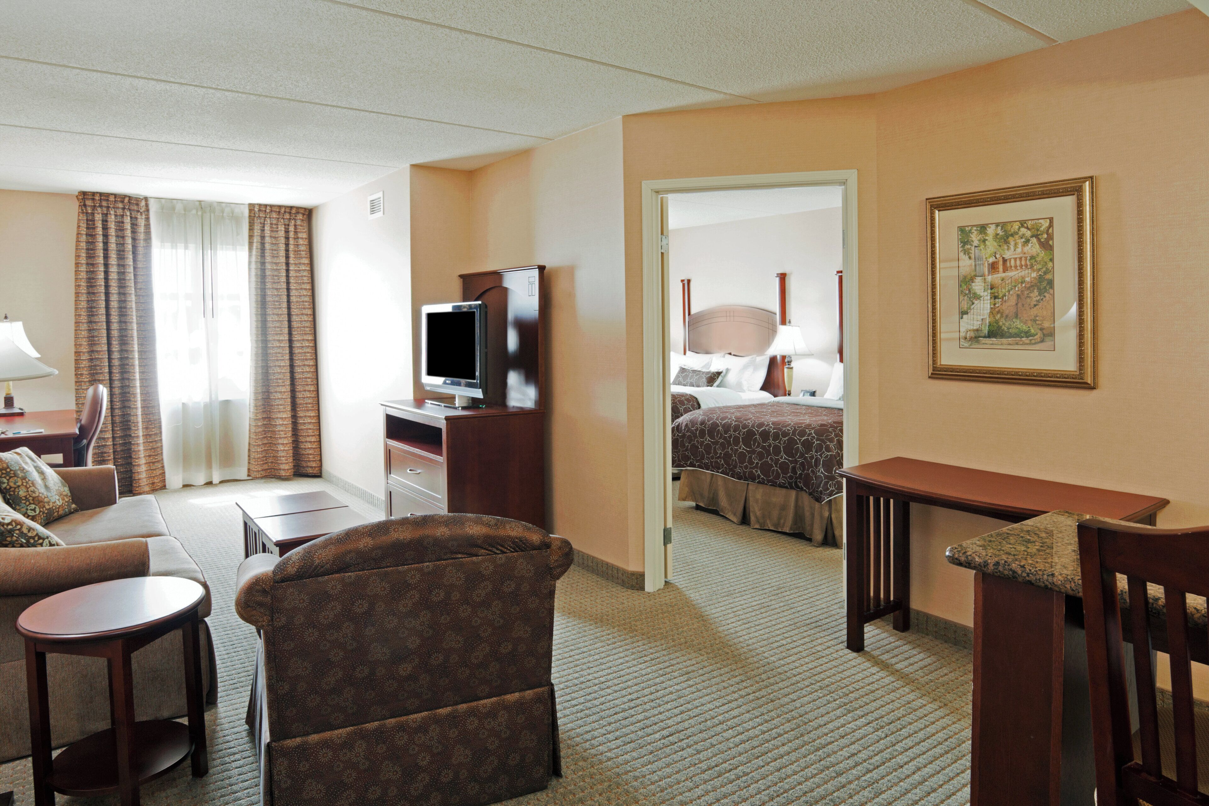 Quarto de hotel com cama, escrivaninha com televisão, sofá, poltrona e pequena mesa.
