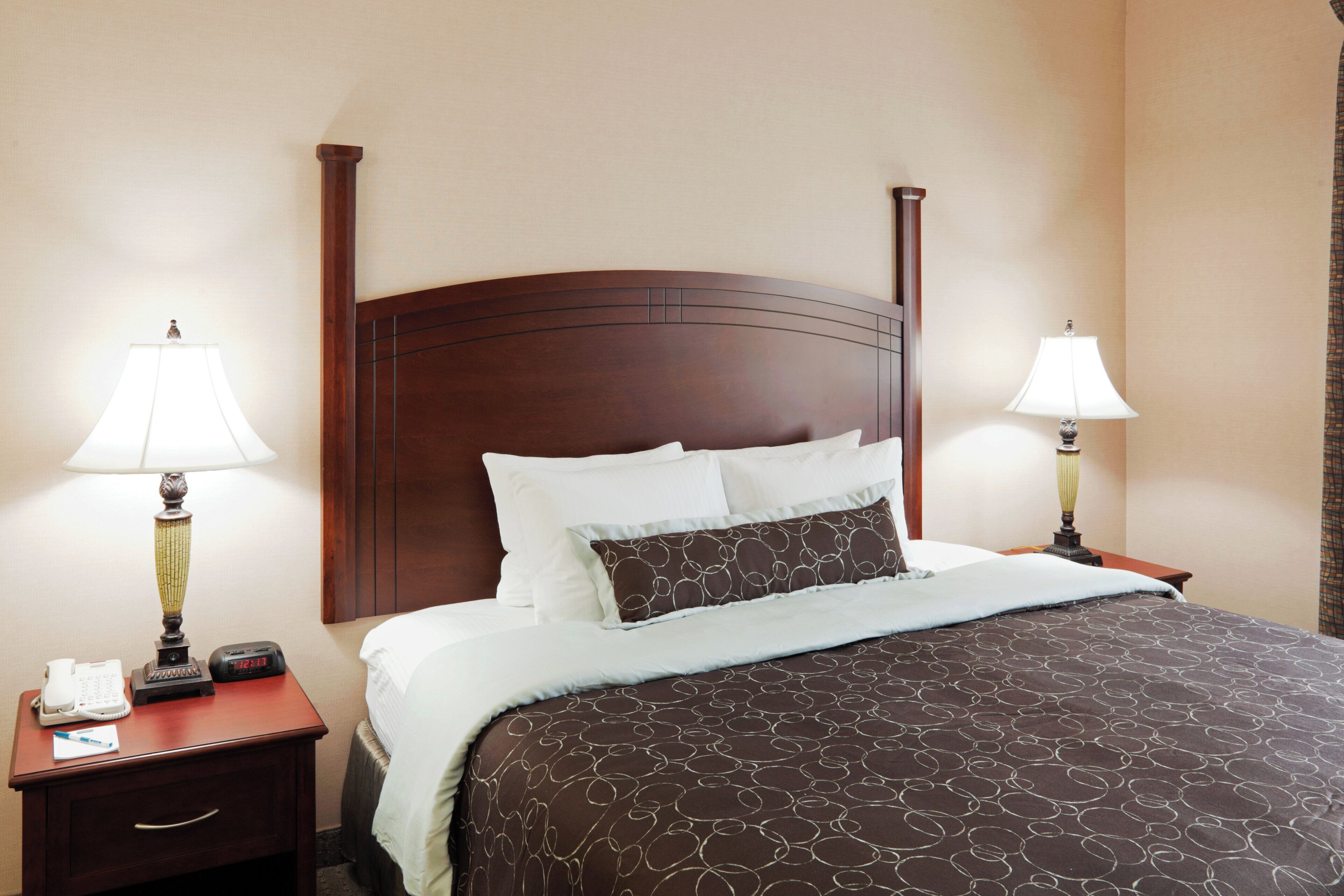 A neatly made bed with a patterned bedspread, two bedside lamps, a nightstand with a telephone and a clock, and a wooden headboard.