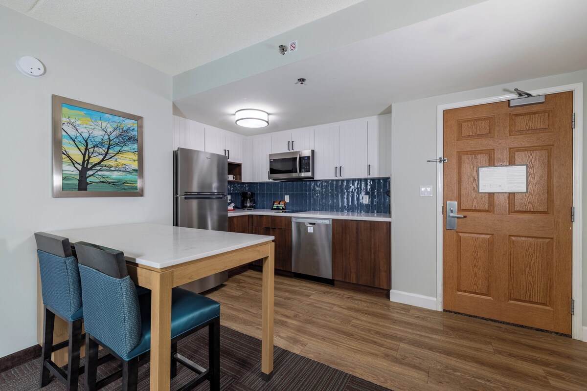 A compact kitchen with white cabinets, a stainless steel refrigerator, a microwave, and a dishwasher. There is a wooden door with a sign, a painting of a tree on the wall, and a table with two chairs.