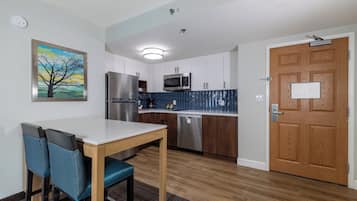 Cozinha compacta com armários brancos, geladeira de aço inoxidável, micro-ondas e lava-louças. Há uma porta de madeira com uma placa, um quadro de uma árvore na parede e uma mesa com duas cadeiras.