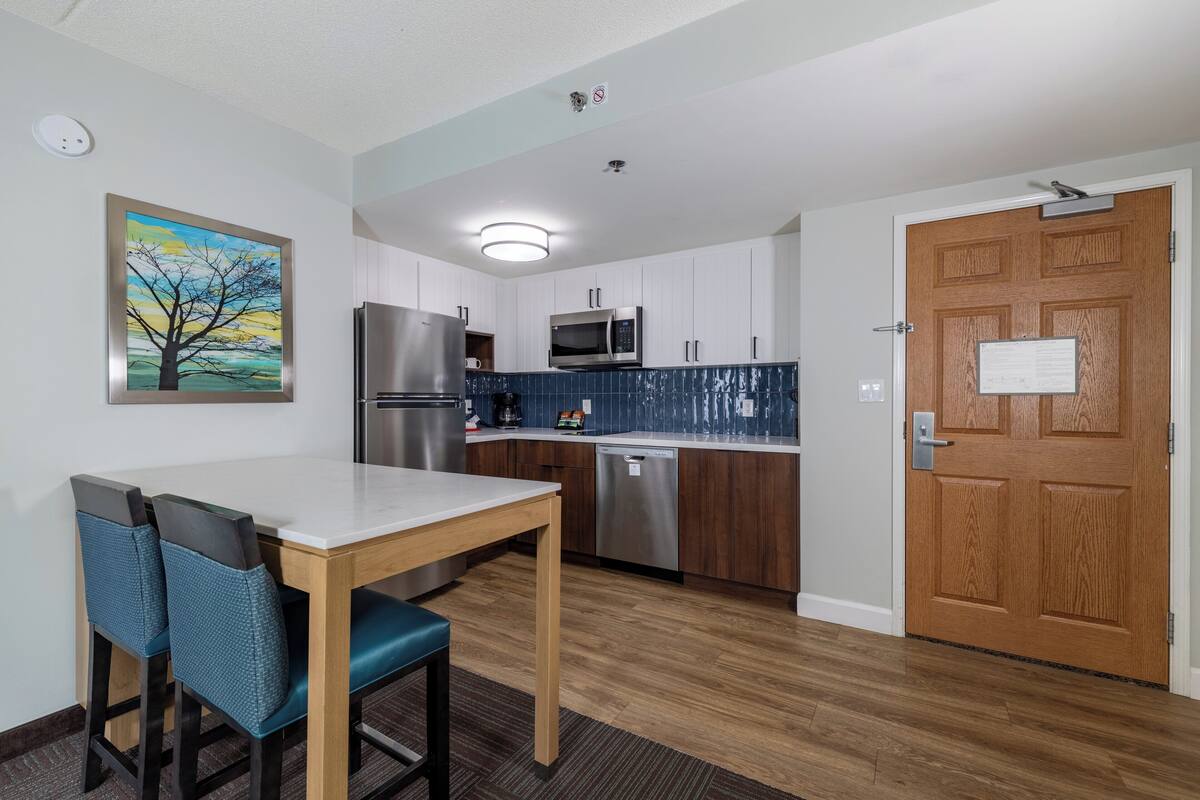 A compact kitchen with white cabinets, a stainless steel refrigerator, a microwave, and a dishwasher. There is a wooden door with a sign, a painting of a tree on the wall, and a table with two chairs.