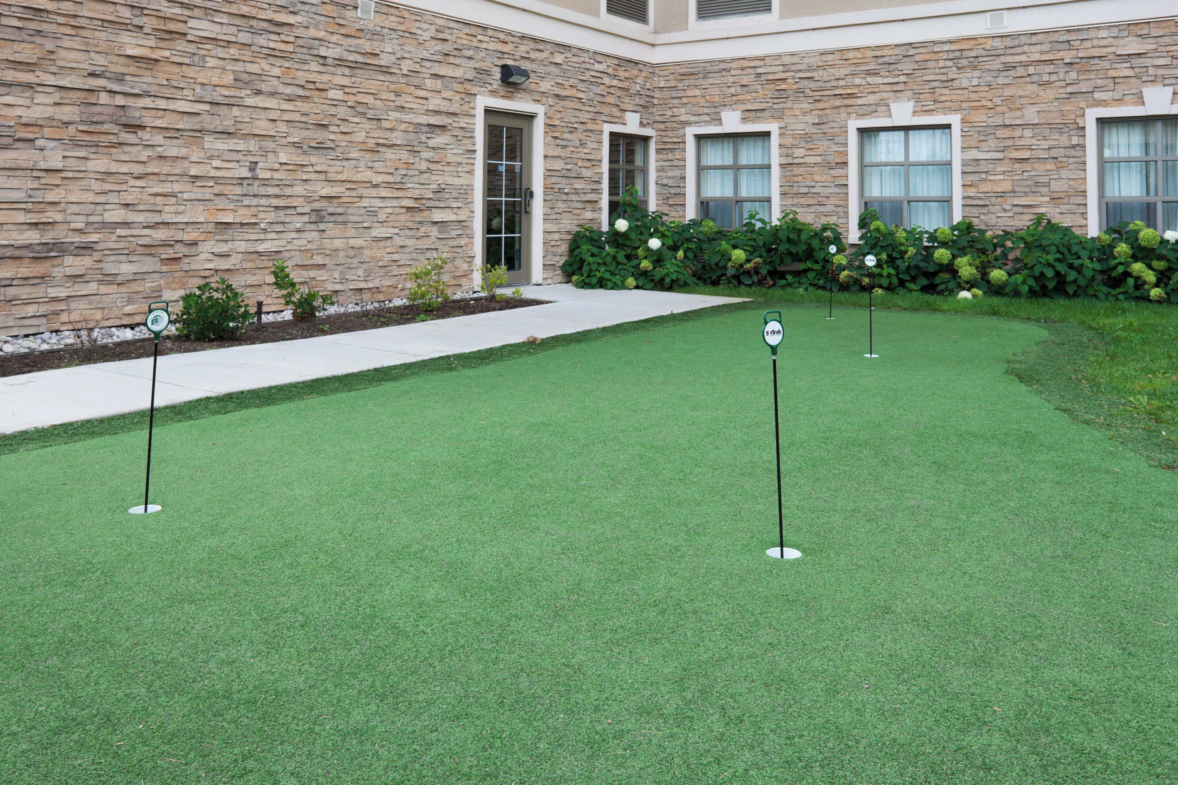 A well-maintained artificial green lawn with golf course markers, a stone building with a door and windows, and a hedge with white flowers.