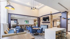 Een ruime hotellobby met een bar in het midden, diverse zithoeken zoals banken en stoelen, een stenen openhaard en een flatscreen televisie aan de muur.