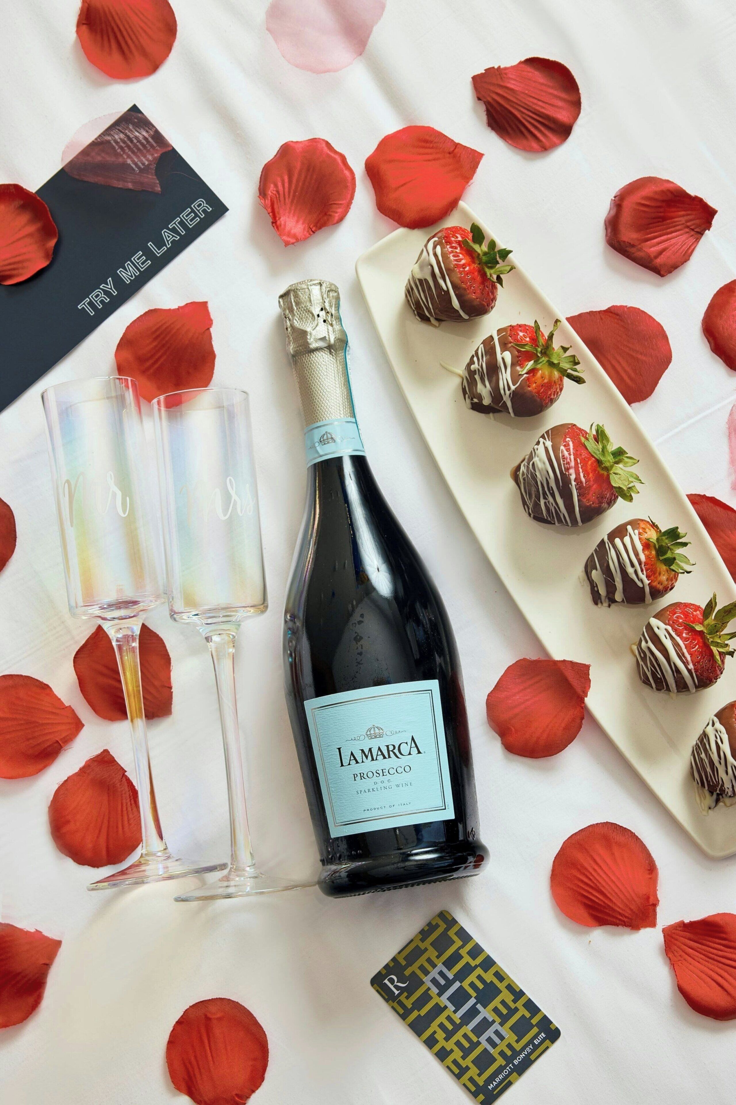 Una botella de vino, dos copas, una tarjeta y fresas bañadas en chocolate sobre una mesa con pétalos de rosa.