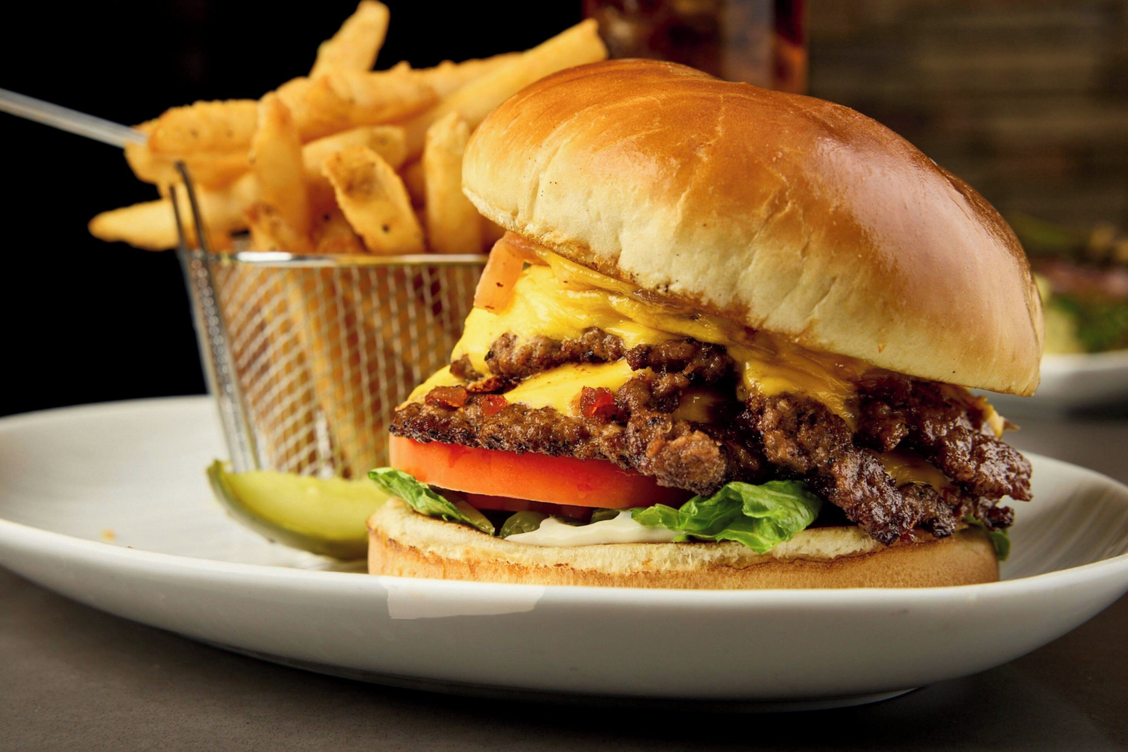 A burger with cheese, beef, and tomato on a bun, accompanied by fries and a pickle.