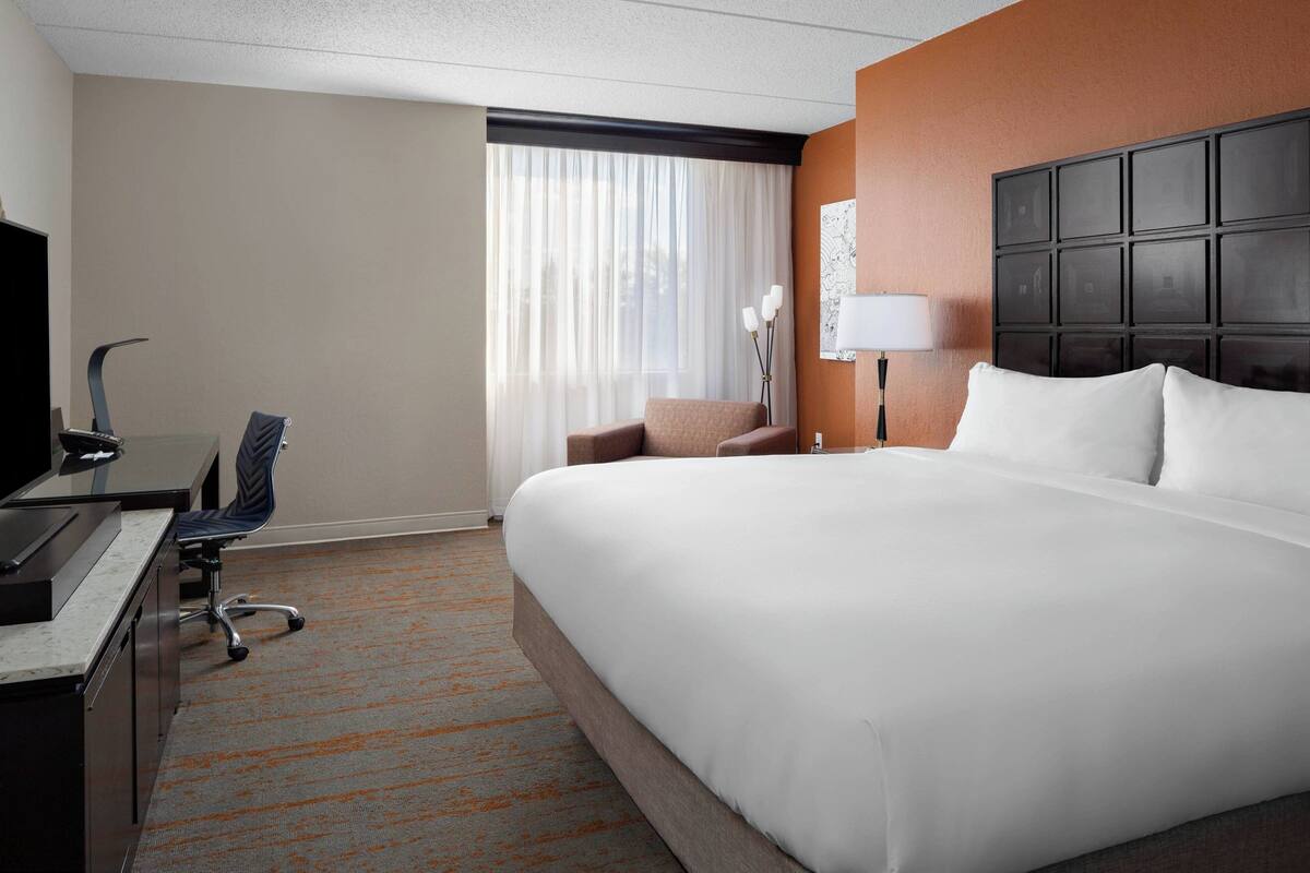 A hotel room with a large bed, a desk with a computer, a chair, a lamp, and a window with curtains.