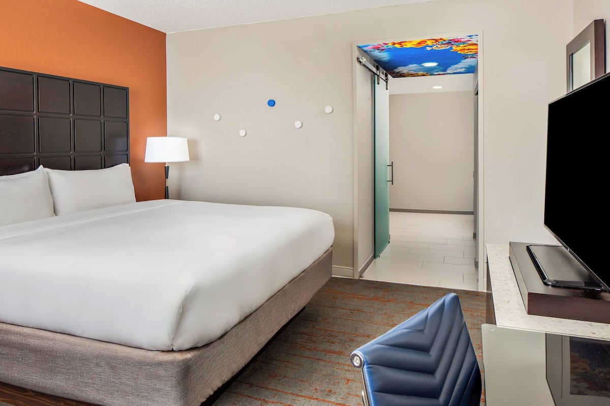 A hotel room with a large bed, a desk with a flat-screen TV, a chair, and a door leading to a bathroom.