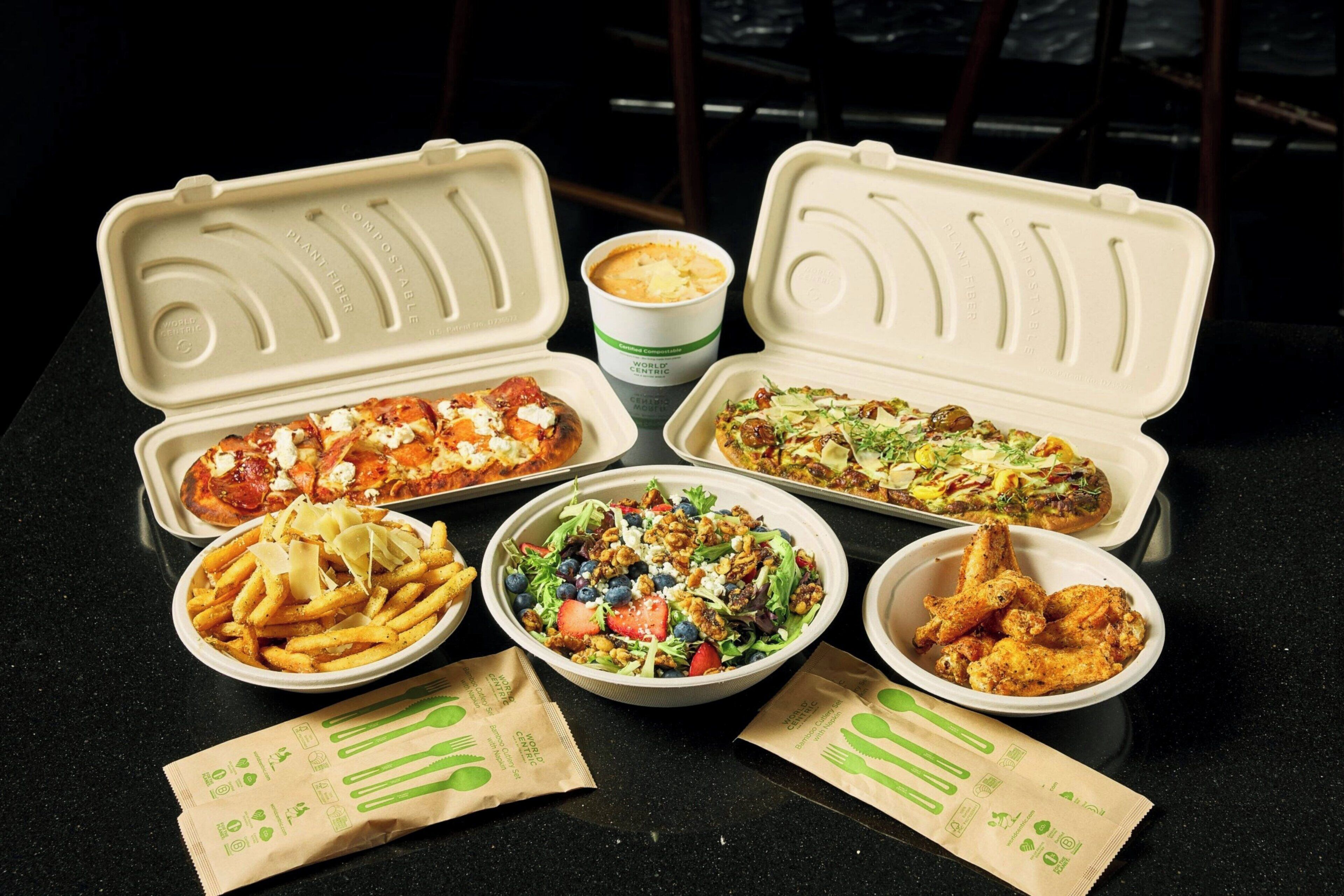 A table with various food items in disposable containers, including pizza, salad, fries, and chicken wings.