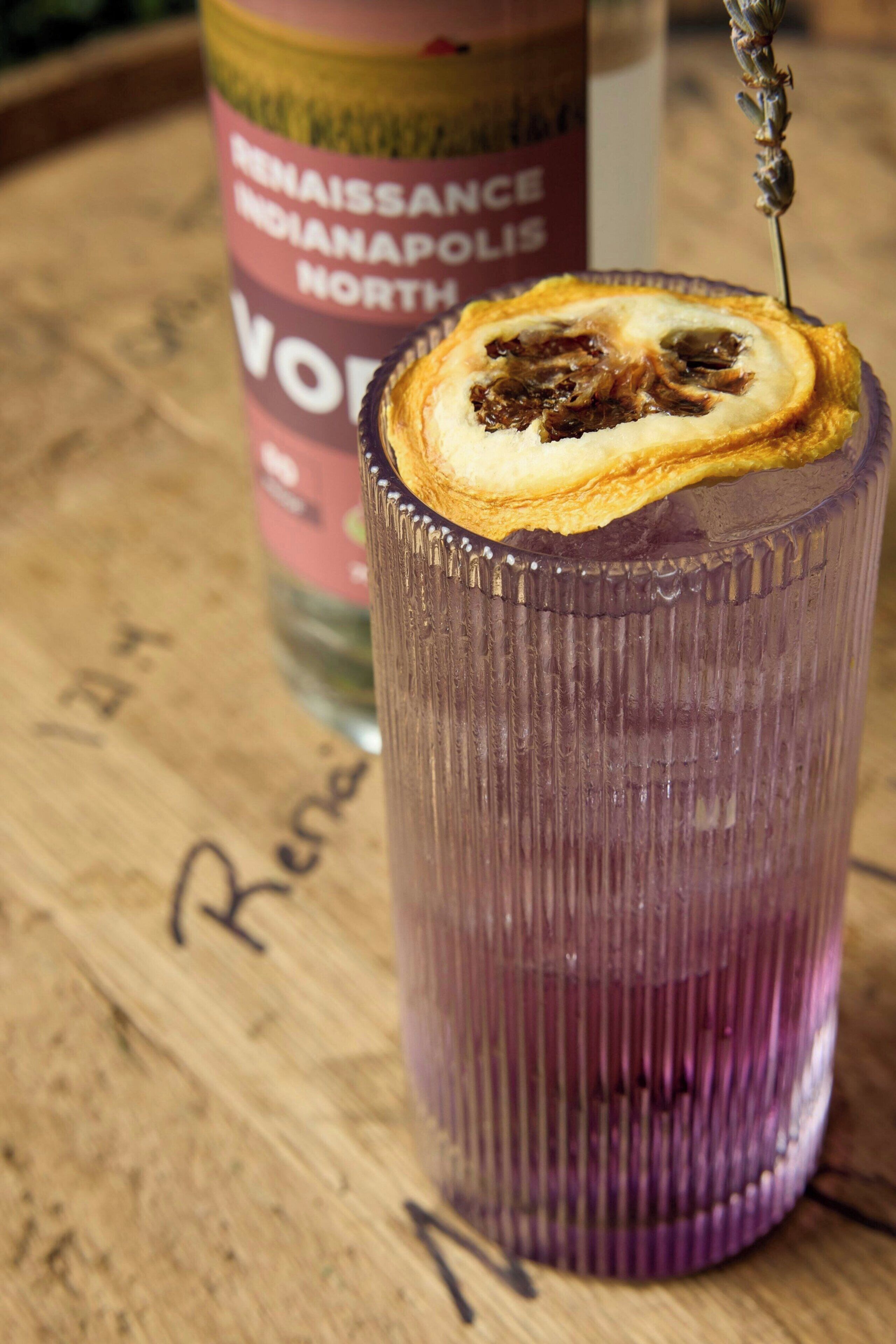 A glass with a slice of fruit on top, a bottle in the background, and a wooden surface with writing.