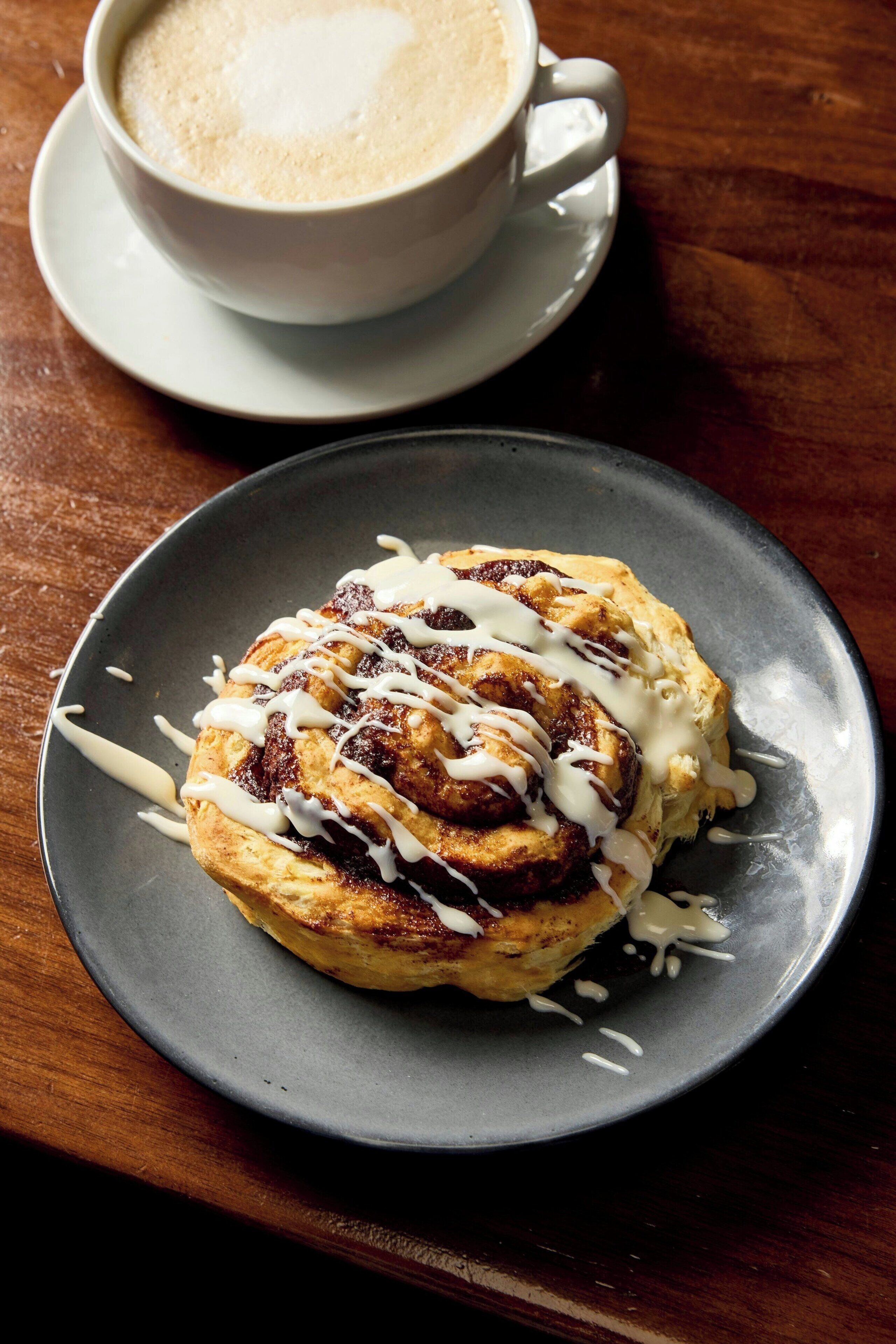 A plate with a cinnamon roll topped with white icing, a cup of coffee, and a saucer on a wooden table.