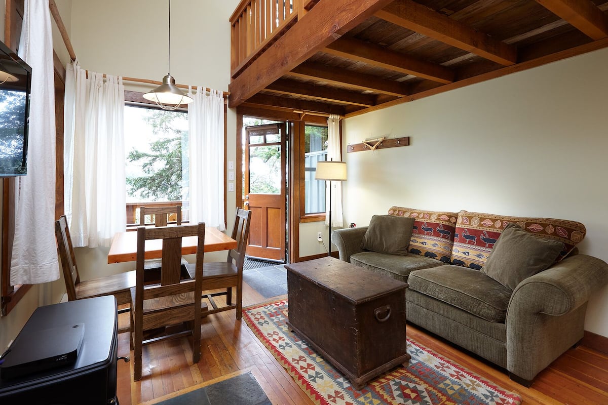 A cozy living room with a wooden ceiling, a sofa with patterned cushions, a wooden coffee table, and a dining area with a table and chairs.