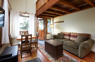 A cozy living room with a wooden ceiling, a sofa with patterned cushions, a wooden coffee table, and a dining area with a table and chairs.